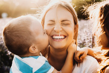 Beautiful Young Mother Laughing With Closed Eyes While Her Son Is Embracing Her Neck And Kissing Her Cheek Outdoor Against Sunset.
