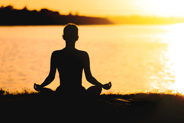 Wall Mural - Beautiful young woman practicing yoga near river at sunset