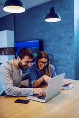 Young cheerful attractive caucasian couple sitting at dining table and paying bills online. Man typing on laptop while woman holding bill.