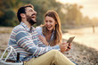 © dusanpetkovic1 - happy laughing caucasian fashionable young couple sitting on coast near river and using tablet. Man holding tablet. Next to man is picnic basket.