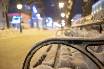 Fragment of a bench covered with snow on a night boulevard in the city in the light of lanterns. Winter cityscape.