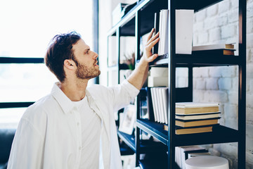 Serious caucasian man in casual wear checking books on home library shelves in living room with modern interior, intelligent hipster guy choosing literature on bookcase learning in apartment.