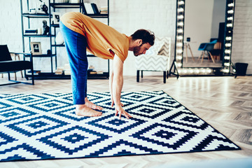 Fototapete - Concentrated young man holding hands on carpet doing morning yoga at home interior.Hipster guy practicing active lifestyle and doing sport exercises during workout in modern flat with stylish
