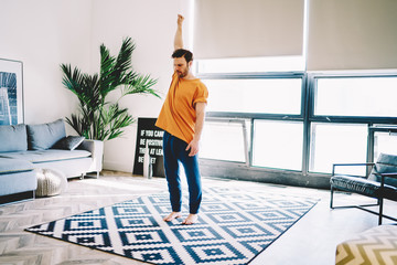 Fototapete - Young man holding hand up and warming up while doing morning exercises on stylish carpet in modern room.Hipster guy practicing yoga during workout at home interior lead a healthy lifestyle