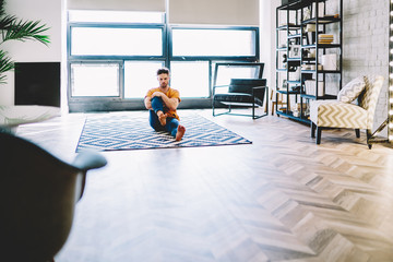 Fototapete - Physically trained young guy stretching leg sitting on cozy carpet in modern comfortable apartment during morning workout.Hipster guy doing yoga exercises to support a healthy lifestyle at home