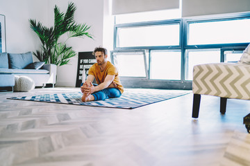 Fototapete - Young man warming up foot before yoga training sitting on carpet in modern apartment with stylish interior.Motivated hipster guy with sport goal stretching leg during workout at home in flat