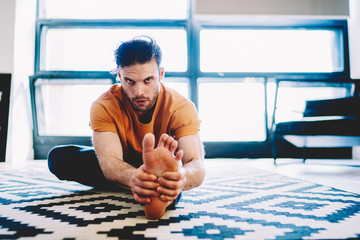 Fototapete - Portrait of motivated and concentrated young man looking at camera while stretching foot and doing morning exercises during yoga training on cozy carpet in modern apartment with stylish interior