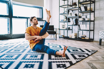 Fototapete - Young man stretching leg and looking up doing sportive exercises during morning workout in modern apartment with home interior,Hipster guy develop flexibility and doing yoga on comfortable carpet