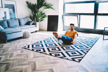 Fototapete - Young man sitting on carpet in modern apartment and doing morning exercises during yoga at home interior.Concentrated hipster guy holding legs and hands up straining the press during workout