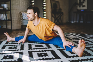 Fototapete - Concentrated young man sitting in twine doing stretching exercises during workout at home interior.Experienced male lover of yoga practicing poses for develop of flexibility in modern apartment