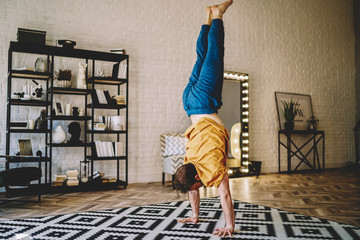 Fototapete - Motivated young man standing on his hands doing morning exercises during yoga at home interior.Male lover of healthy lifestyle develop strength of hands practicing active training in apartment
