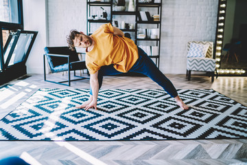Fototapete - Physically trained young man in sportive wear doing stretching exercises during morning workout on comfortable carpet in home apartment.Motivated hipster guy warming up before yoga training