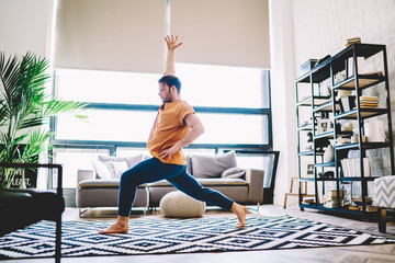 Fototapete - Side view of sportive young man holding hand up during morning workout at home apartment.Hipster guy warming up before yoga training on stylish carpet in flat enjoying harmony and healthy vitality