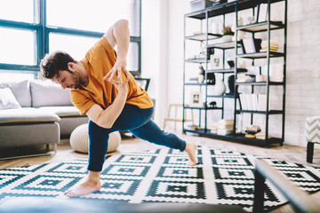Fototapete - Motivated flexible young man in sportive wear engaged yoga training on stylish carpet in modern apartment in morning free time.Male lover of meditation standing in sport pose during workout