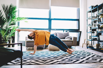 Fototapete - Experienced male lover of yoga in sportive wear stretching hand standing in yoga pose during morning training at home interior.Professional sportive man practising meditation in modern apartment