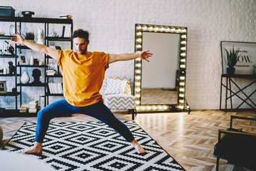Fototapete - Professional young man standing on carpet and raising hands on sides during yoga and meditation in morning time at home interior.Concentrated hipster guy practicing relaxation in apartment