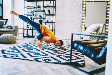 Fototapete - Portrait of positive professional young man standing on hands in asana pose on cozy carpet in modern apartment.Smiling hipster guy doing yoga exercises during morning training at home