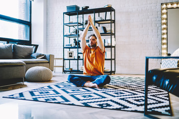 Fototapete - Calm young bearded man sitting in lotus pose on cozy carpet and holding hands in namaste above head during meditation at home apartment.Hipster guy with closed eyes doing yoga exercises