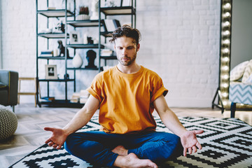Fototapete - Concentrated young bearded man in active wear sitting in lotus pose during morning training on comfortable carpet in modern apartment.Calm hipster guy engaged in yoga at home interior