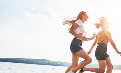 Wall Mural - Two female friends runs and have fun at beach near the lake at sunny daytime