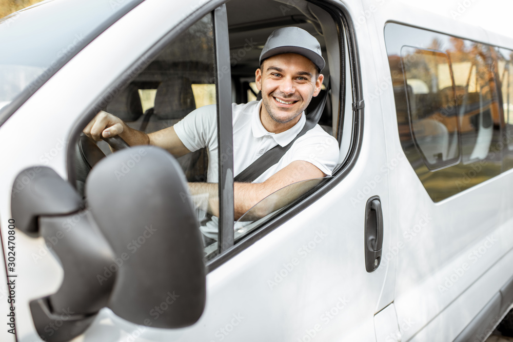 Portrait of a cheerful delivery driver in uniform looking out the window of the white cargo van vahicle, delivering goods by car - obrazy, fototapety, plakaty 