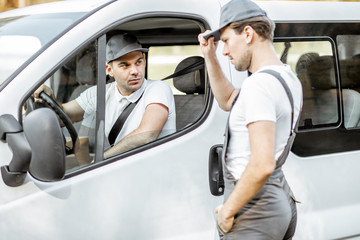 Delivery company employees in uniform delivering goods by cargo van vehicle, mover talking with a car driver