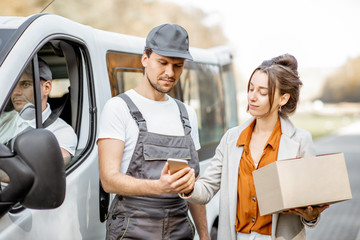 Delivery company employees in uniform delivering goods to a client by cargo van vehicle, woman signing on a smartphone, receiving parcel outdoors