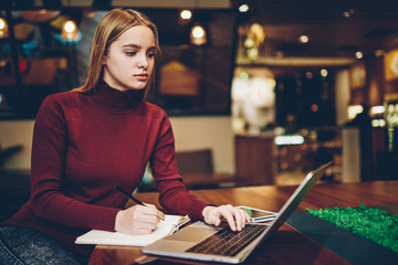 Canvas Print - Young pretty clever female copywriter watching training webinar in website on laptop and recording useful information to notepad.Intelligent hipster girl reading publication in web page indoors