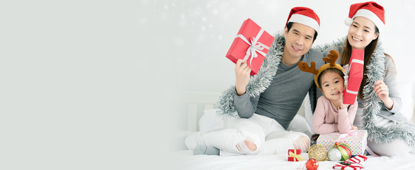 Wall Mural - Asian family have father, mother and daughter with gray, pink casual wear and Santa hats. Sitting on a white bed with a red gift box And  small ball. Celebrate Christmas and Happy New Year. copy space