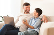 © Prostock-studio - Father And Son Using Phone And Laptop Sitting At Home