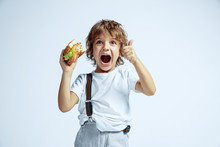 Pretty Young Curly Boy In Casual Clothes On White Studio Background. Eating Burger. Caucasian Male Preschooler With Bright Facial Emotions. Childhood, Expression, Fun, Fast Food. Showing Thumb Up.