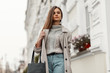 © alones - Elegant young woman is walking in a stylish trench coat with a black handbag in a warm knitted sweater near vintage buildings with flowers on the facades.Attractive lady enjoys a walk on a city street