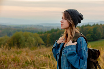 Wall Mural - Style girl in denim jacket and hat with backpack in countryside with mountains on background