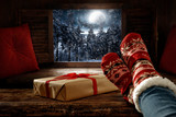 Fototapeta  - Wooden winter background of free space.Christmas december time.Woman legs with woolen socks and night landscape of forest with mountains and big moon on sky.Retro old frame and pillows decoration.