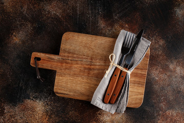 Cutlery on the plate on dark brown background. Fork and table-knife with wooden handles on linen napkin and wooden board