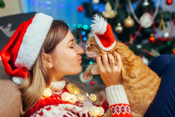 Christmas celebration with cat. Woman playing and kissing pet in Santa's hat by New year tree at home.