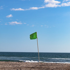 Green flag, beach scene in a sunny bright day. Blue sky with white clouds. 1x1.