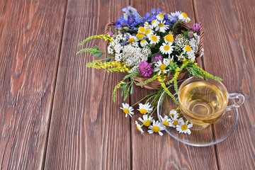 Herbal tea with the chamomile flowers in a glass cup and fresh wild flowers in a basket on a wooden background.