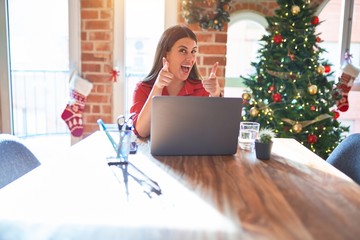 Poster - Beautiful woman sitting at the table working with laptop at home around christmas tree pointing fingers to camera with happy and funny face. Good energy and vibes.