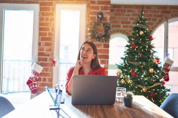 Sticker - Beautiful woman sitting at the table working with laptop at home around christmas tree with hand on chin thinking about question, pensive expression. Smiling and thoughtful face. Doubt concept.