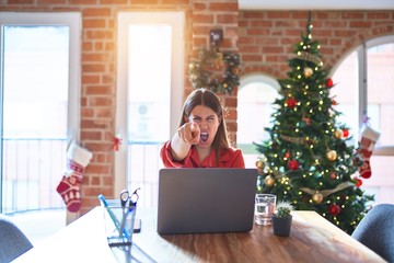 Sticker - Beautiful woman sitting at the table working with laptop at home around christmas tree pointing displeased and frustrated to the camera, angry and furious with you