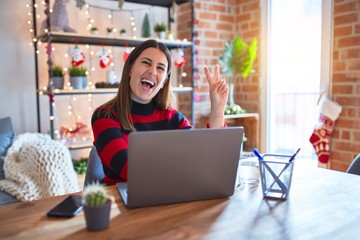 Poster - Beautiful woman sitting at the table working with laptop at home around christmas lights smiling with happy face winking at the camera doing victory sign. Number two.