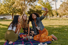 Group of happy diverse three female friends sitting together on blanket over the green grass enjoying at picnic taking selfie on smartphone in the park