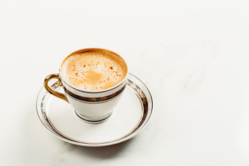 Cup of fresh americano or espresso coffee with golden foam froth on pile of brown raw coffee beans on white marble table background. Morning hot drink, coffee break, cope space