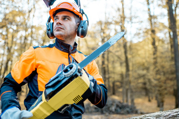 Canvas Print - Portrait of a professional lumberjack in protective workwear standing with a chainsaw on a pile of logs in the forest