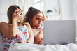 © standret - Watching scary movie. Two female friends in sleepwear lying on the bed and using laptop