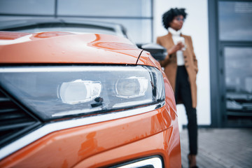 With cup of drink. Young african american woman in glasses stands outdoors near modern car