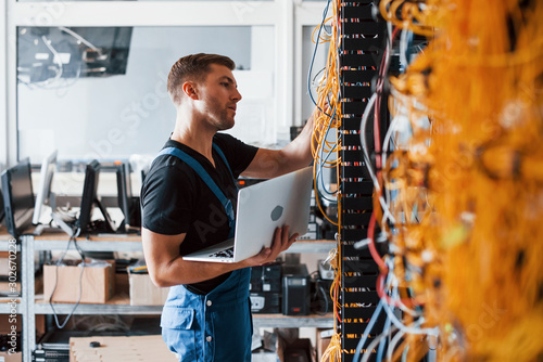 Fotomural Young man in uniform and with laptop works with internet equipment and wires in