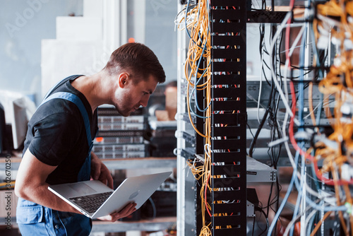 Fotografía Young man in uniform and with laptop works with internet equipment and wires in
