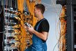Young man in uniform with measuring device works with internet equipment and wires in server room